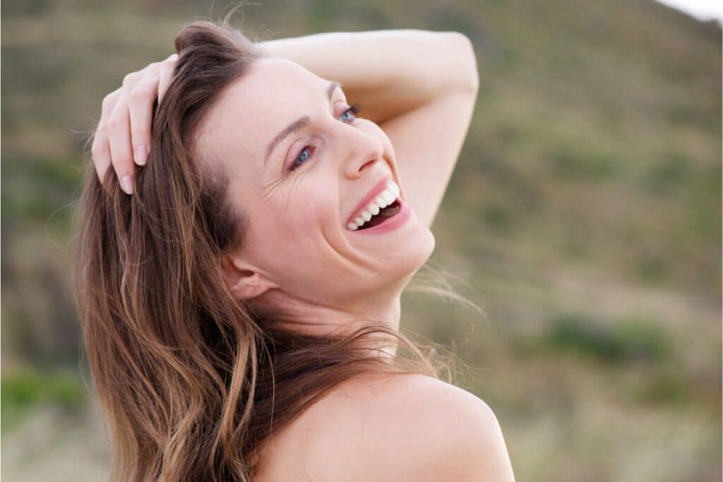 A woman with long brown hair smiles and looks over her shoulder whilst holding one hand in her hair. The blurred outdoor background, filled with green and brown tones, evokes a sense of calm and natural health benefits.