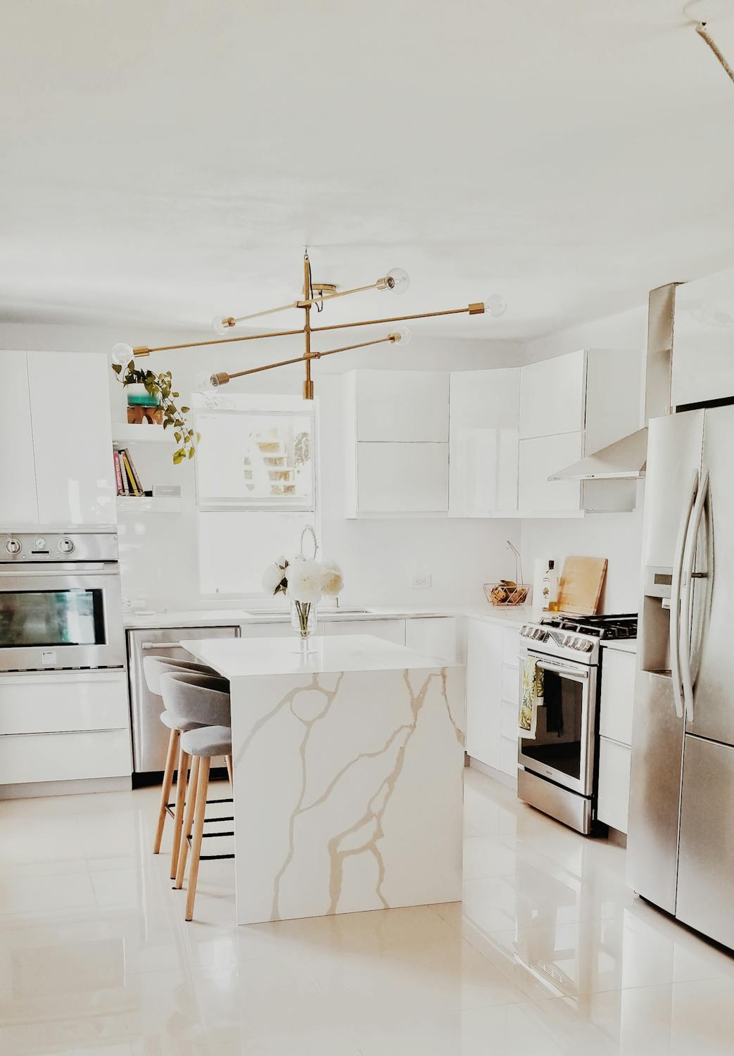 A modern white kitchen features glossy cupboards, stainless steel appliances, an island with marble-patterned sides, grey barstools, a gold chandelier, and a vase of flowers on the worktop for a fresh kitchen ambience.
