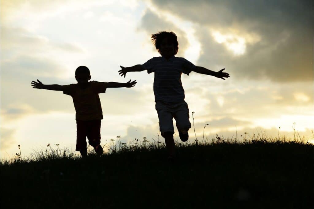 Two children are running with outstretched arms on a grassy hill at sunset, their silhouettes highlighted against a cloudy sky as they play carefree games.