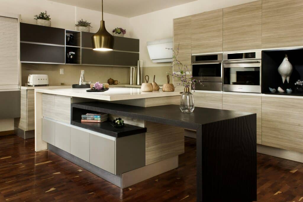 Modern kitchen with light wood cupboards, built-in appliances, a white worktop island with a black extension, pendant lighting, and stylish kitchen décor including a vase with flowers and wooden chopping boards.