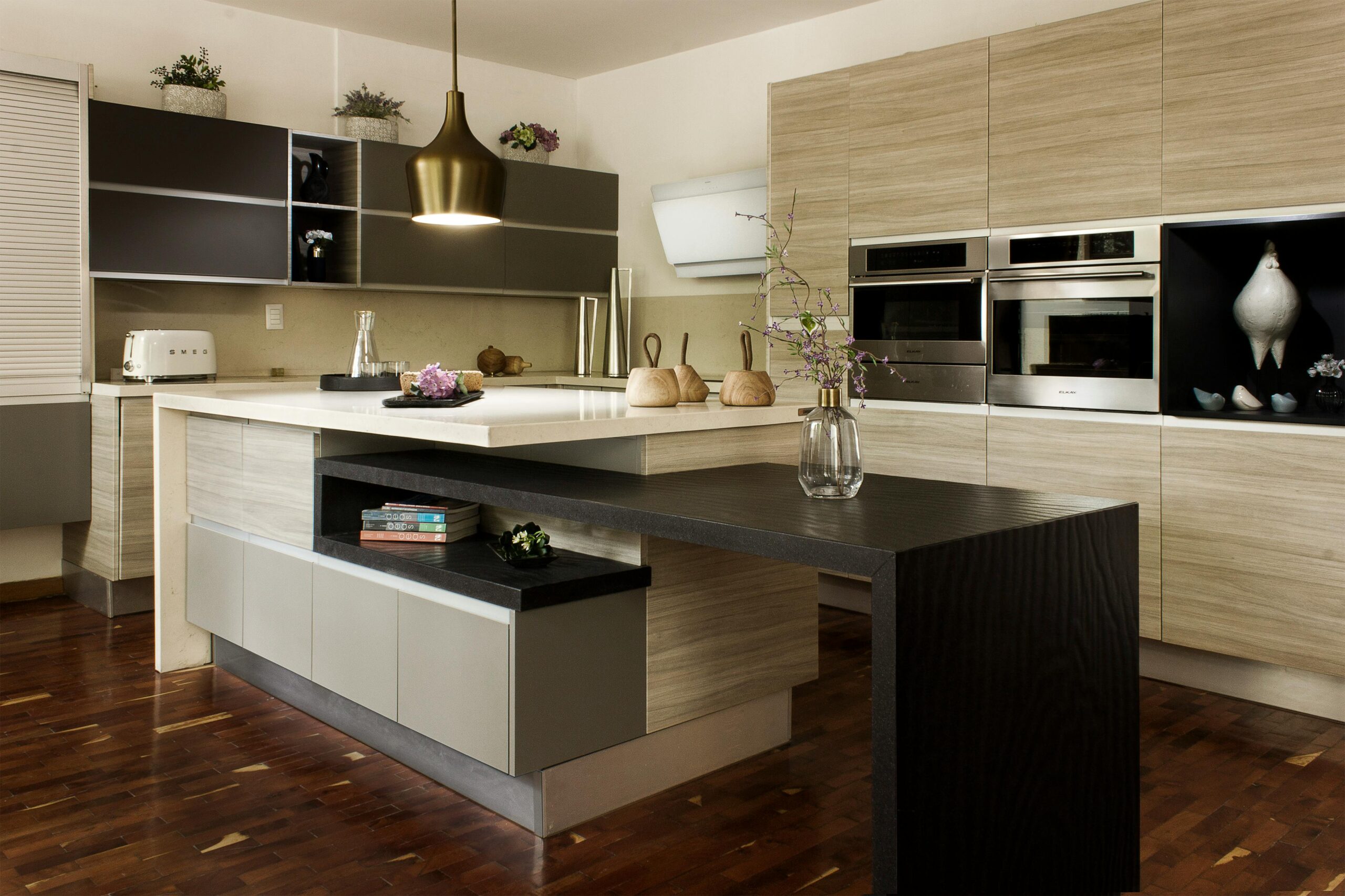Modern kitchen with light wood cupboards, built-in appliances, a white worktop island with a black extension, pendant lighting, and stylish kitchen décor including a vase with flowers and wooden chopping boards.