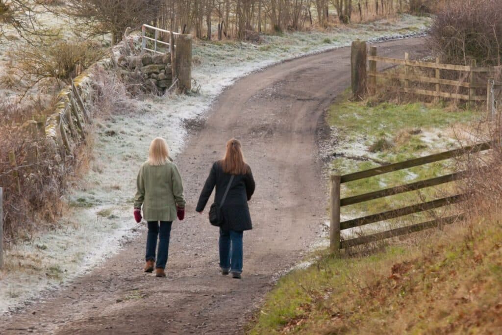 Two women in winter coats walk along a winding, frosty dirt path bordered by wooden fences and lightly frosted grass, surrounded by bare trees on a chilly day.