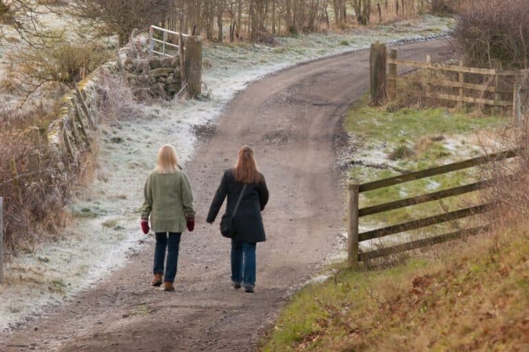 Two women in winter coats walk along a winding, frosty dirt path bordered by wooden fences and lightly frosted grass, surrounded by bare trees on a chilly day.