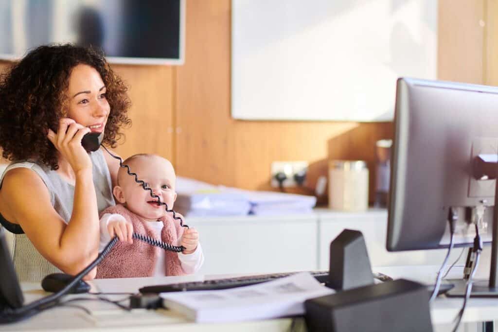 A businesswoman sits at an office desk, talking on the phone whilst holding a smiling baby in her lap. The baby is grabbing the phone cord. A computer monitor and papers are on the desk.