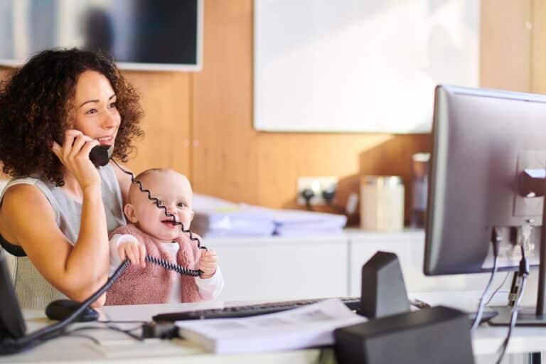 A businesswoman sits at an office desk, talking on the phone whilst holding a smiling baby in her lap. The baby is grabbing the phone cord. A computer monitor and papers are on the desk.
