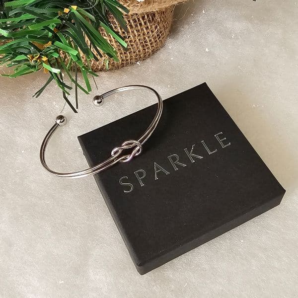 A silver knot bangle bracelet rests on a closed black box with "SPARKLE" printed on top, next to green artificial foliage wrapped in hessian—a perfect piece for any gift guide.