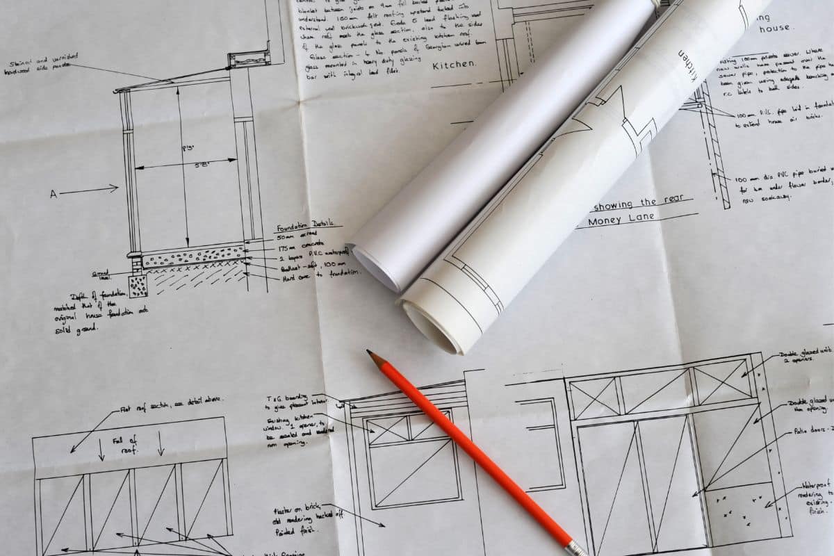 Two rolled-up architectural blueprints rest on an open garden blueprint with technical drawings and handwritten notes. A sharpened orange pencil lies nearby. The blueprints appear to detail building construction plans and landscaping ideas.