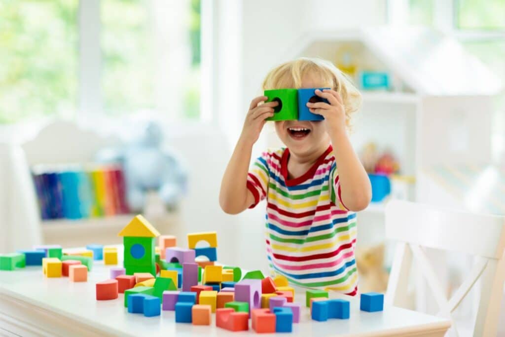 A young child in a rainbow-striped shirt smiles and holds two colourful educational toys to their eyes, sitting at a table covered with various building bricks in a bright, playful room.