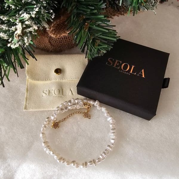 A pearl bracelet with a gold clasp is displayed near a SEOLA-branded black box and a cream pouch. Set on a snowy surface with a pine branch, it’s an elegant addition to any holiday gift guide.