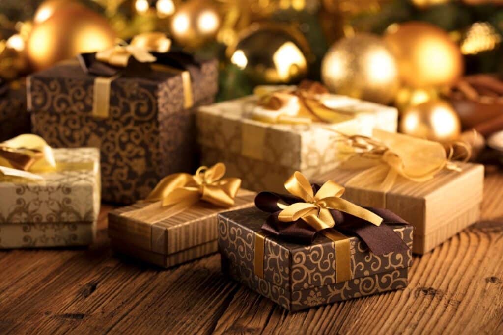 A group of elegant gift boxes with gold and brown wrapping paper and ribbon bows sits on a wooden surface, with blurred golden ornaments in the background.