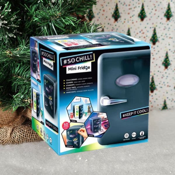 A boxed "#SO CHILL! Mini Fridge" sits on a surface next to a decorated Christmas tree, set against a festive backdrop with small tree patterns—perfect for any holiday gift guide.