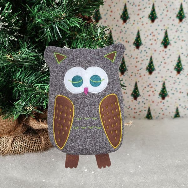 A handmade felt owl ornament with green eyes stands on a snowy surface near a decorated Christmas tree, making it a charming addition to any holiday gift guide. The festive background features small, repeating Christmas tree patterns.