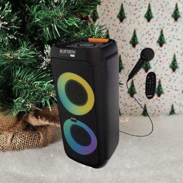 A portable Bluetooth speaker with colourful LED lights, a microphone, and a remote control sits next to a decorated Christmas tree—an ideal choice for any holiday gift guide, set against a festive backdrop with small green trees.
