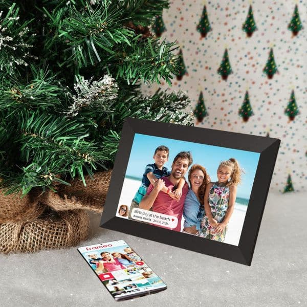 A digital photo frame displaying a family beach picture sits on the floor beside a decorated Christmas tree, featured in a holiday gift guide, with a mobile phone showing photo-sharing app options in front of the frame.