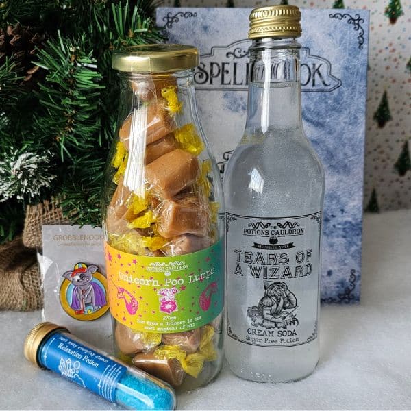 A magical gift guide pick: A glass bottle of caramel treats labelled "Unicorn Poo Lumps," a bottle of "Tears of a Wizard" cream soda, a vial of blue powder, and a wizard-themed card set against a Spellbook backdrop.