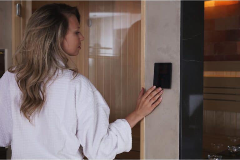 A woman in a white dressing gown stands by a wall, pressing a control panel outside a wooden sauna, preparing to enjoy the soothing warmth within.