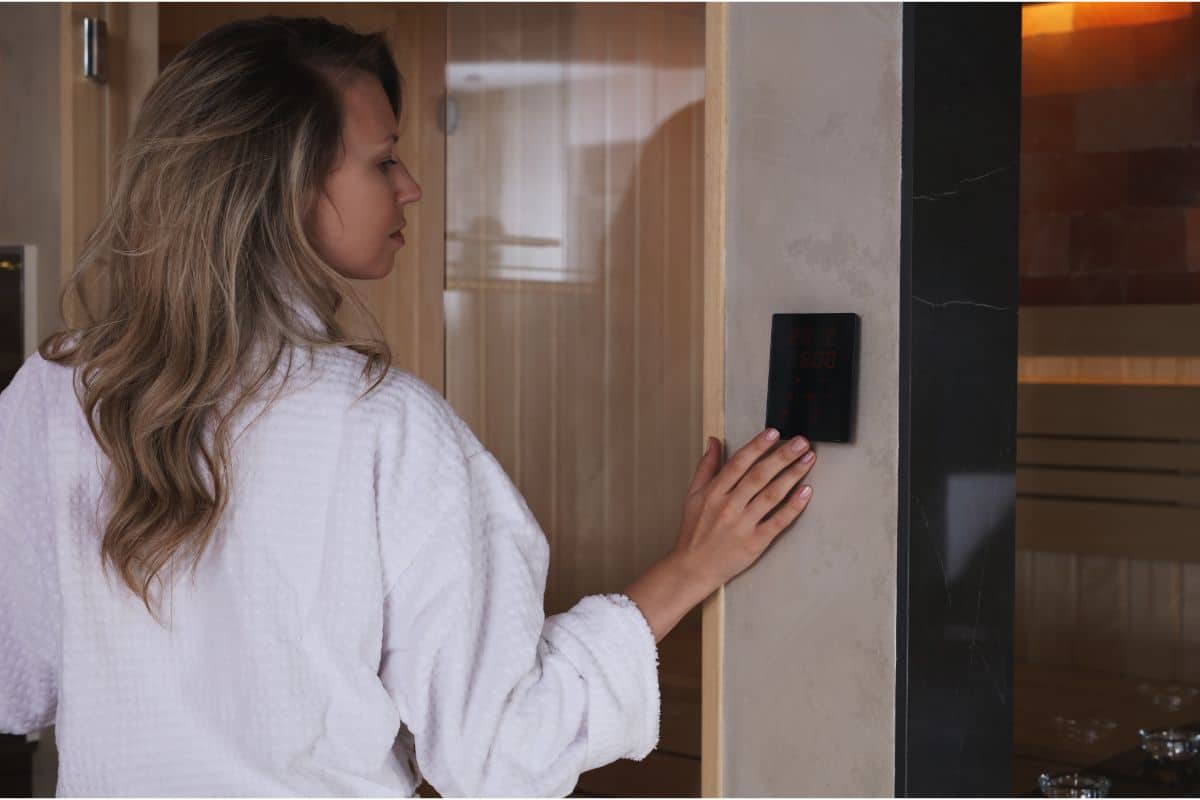 A woman in a white dressing gown stands by a wall, pressing a control panel outside a wooden sauna, preparing to enjoy the soothing warmth within.