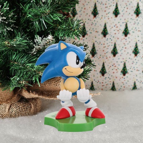 A Sonic the Hedgehog figurine stands on a green base in front of a decorated Christmas tree and festive, tree-patterned wallpaper—perfect for any holiday gift guide.
