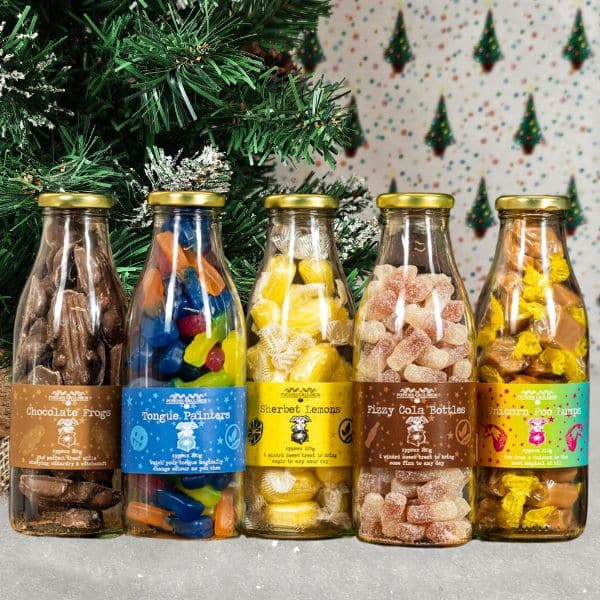 Five glass bottles filled with colourful sweets, including chocolate frogs, gummy sweets, and sherbet lemons, are displayed in front of a Christmas tree—perfect for any holiday gift guide.