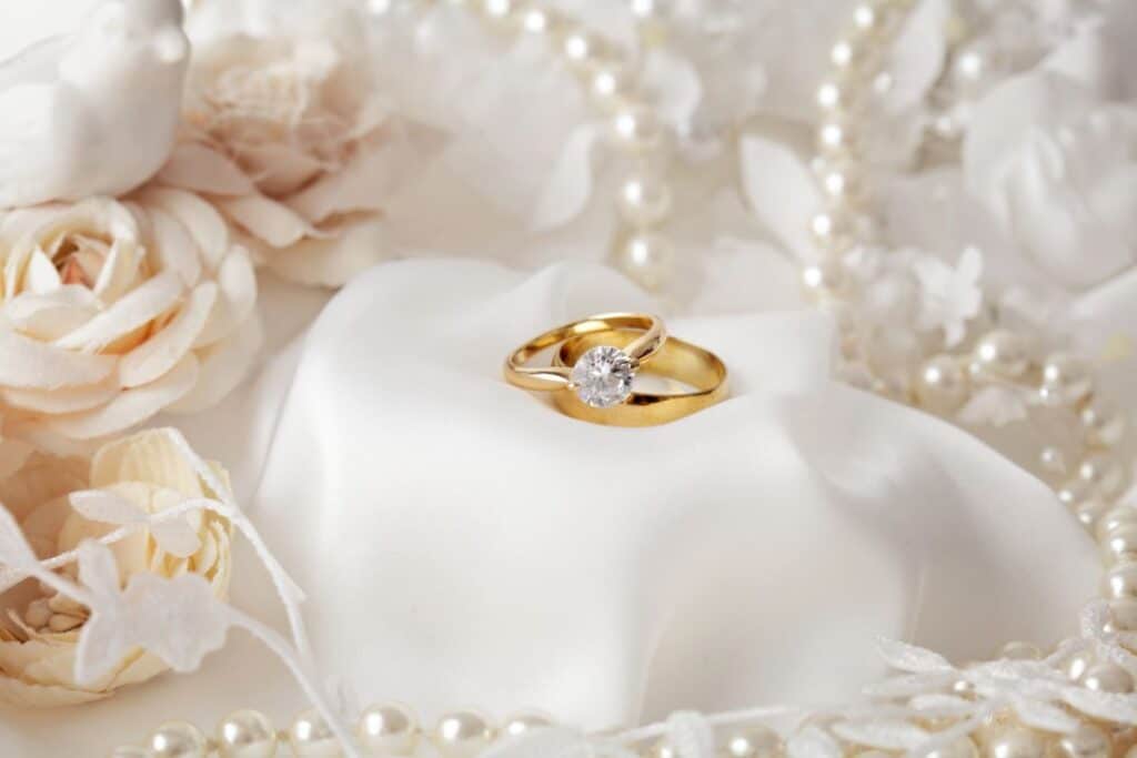 A gold engagement ring with a single diamond is displayed on a white satin cloth, surrounded by soft pink roses, white wedding floral decorations, and pearl beads.