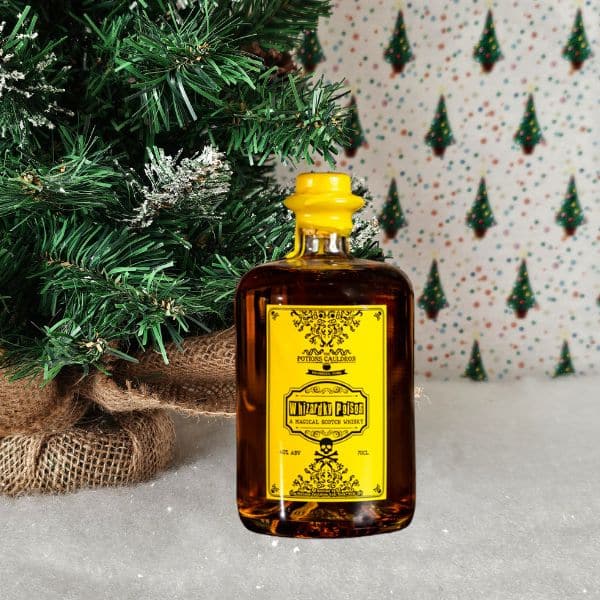 A bottle of whisky with a bright yellow label stands on artificial snow beside a decorated Christmas tree with hessian at its base—a perfect addition to any holiday gift guide. The background features a festive pattern of small Christmas trees.