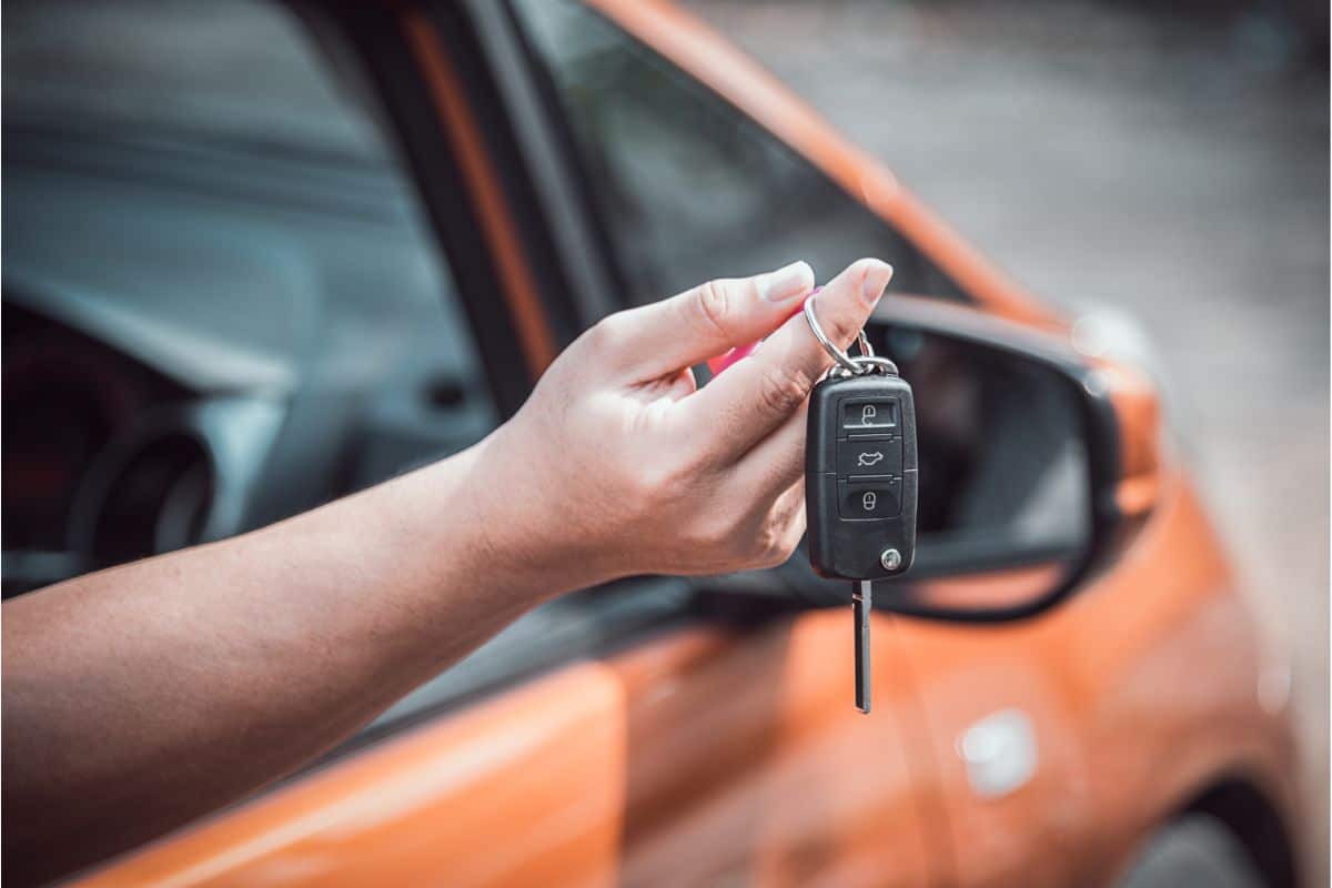 A hand holds a car key fob near an orange used car with the driver-side window open, while the wing mirror is visible in the background.