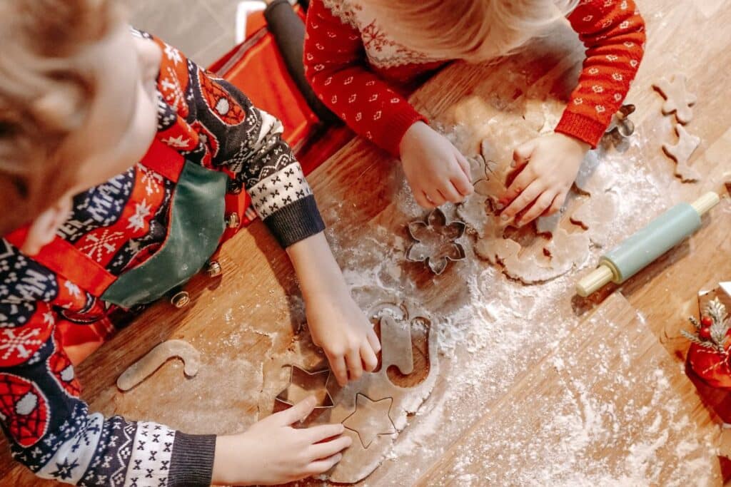 Two children in festive Christmas jumpers use biscuit cutters to shape dough on a floured wooden table, surrounded by baking tools including a rolling pin.