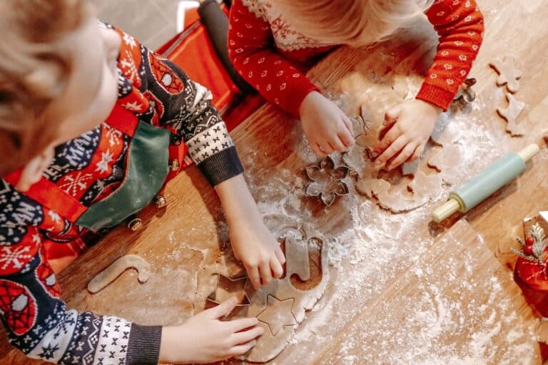Two children in festive Christmas jumpers use biscuit cutters to shape dough on a floured wooden table, surrounded by baking tools including a rolling pin.