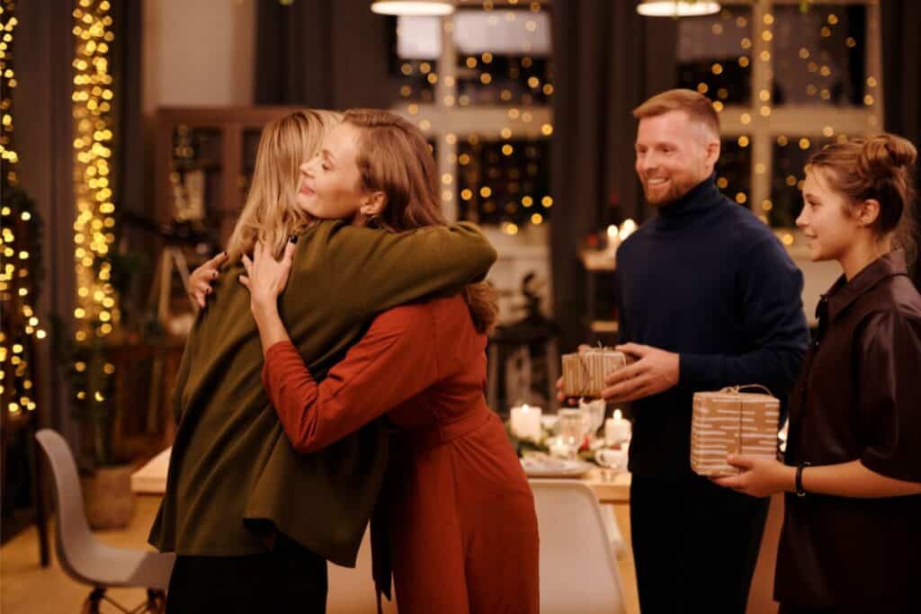 Two women hug and smile at a festive, warmly lit Christmas gathering. A man and a young woman stand nearby, both holding wrapped presents. Twinkling lights and a decorated table are visible in the background.