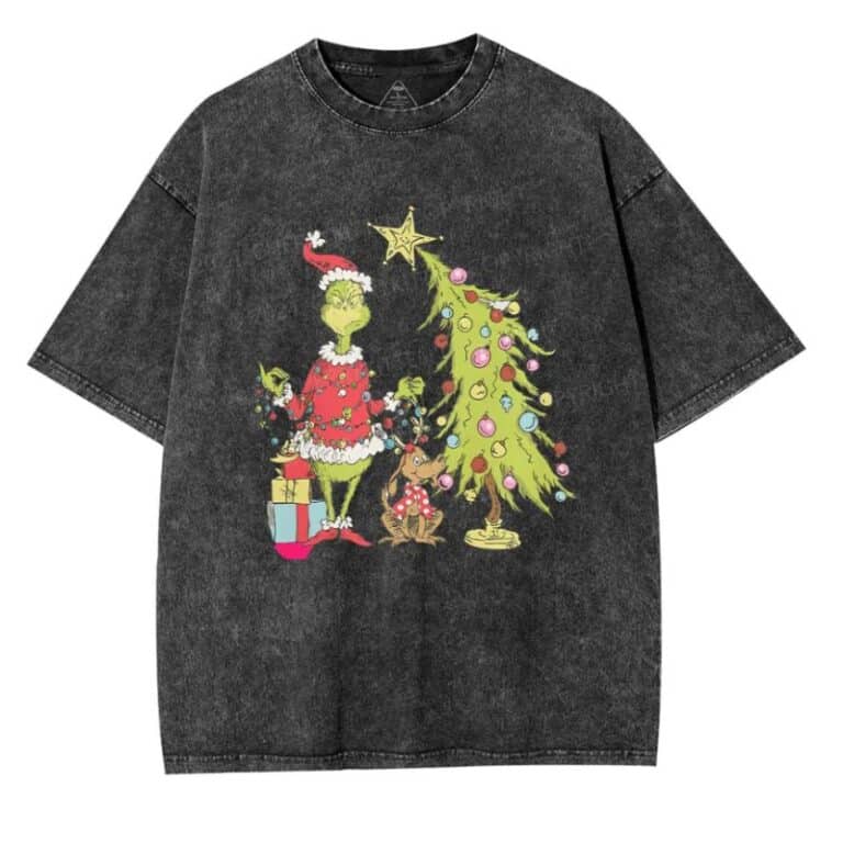 A black T-shirt featuring a festive illustration of the Grinch in a Santa outfit, standing beside a decorated Christmas tree with presents and a small Cherry Kitten wearing reindeer antlers.