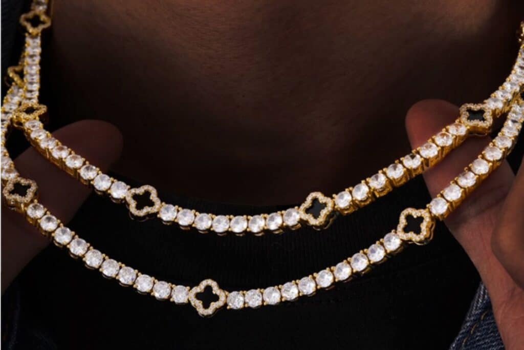 Two hands hold up two gold and diamond jewellery necklaces, each featuring clover-shaped links between the round diamonds, against a dark background.