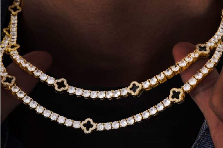Two hands hold up two gold and diamond jewellery necklaces, each featuring clover-shaped links between the round diamonds, against a dark background.