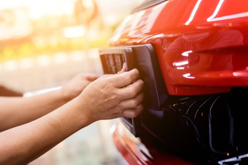 Close-up of a person’s hands attaching or removing private number plates from the rear of a shiny red car. The background is blurred, focusing on the hands and the vehicle.
