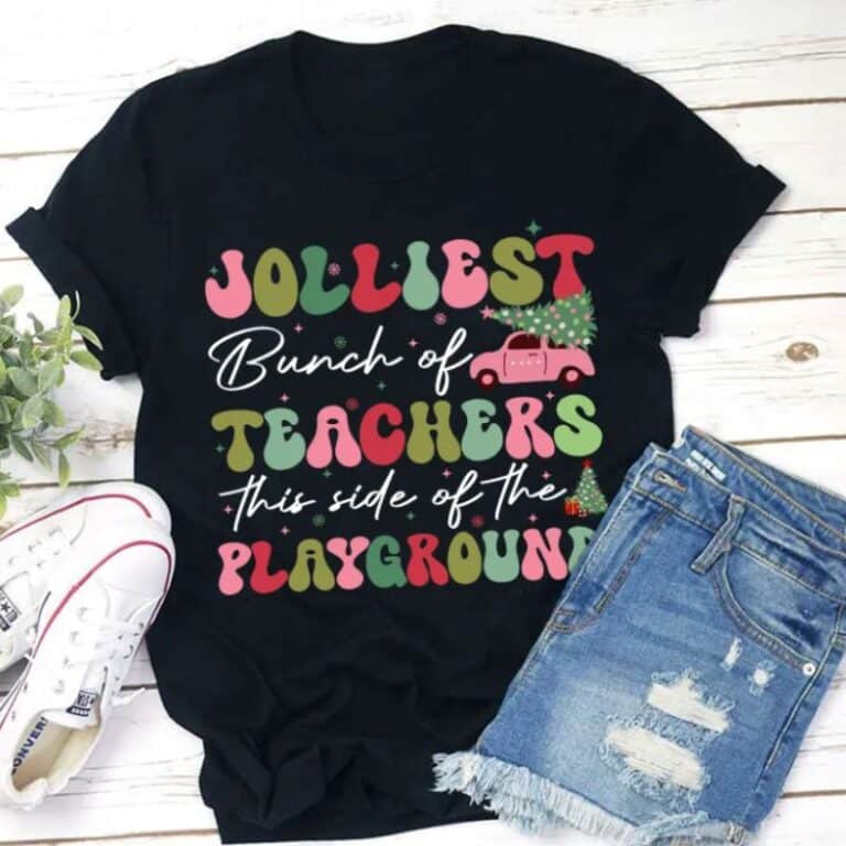A black T-shirt for teachers featuring colourful text, "Jolliest Bunch of Teachers This Side of the Playground," displayed with ripped denim shorts and white trainers. The festive design includes a Christmas tree and a red lorry.