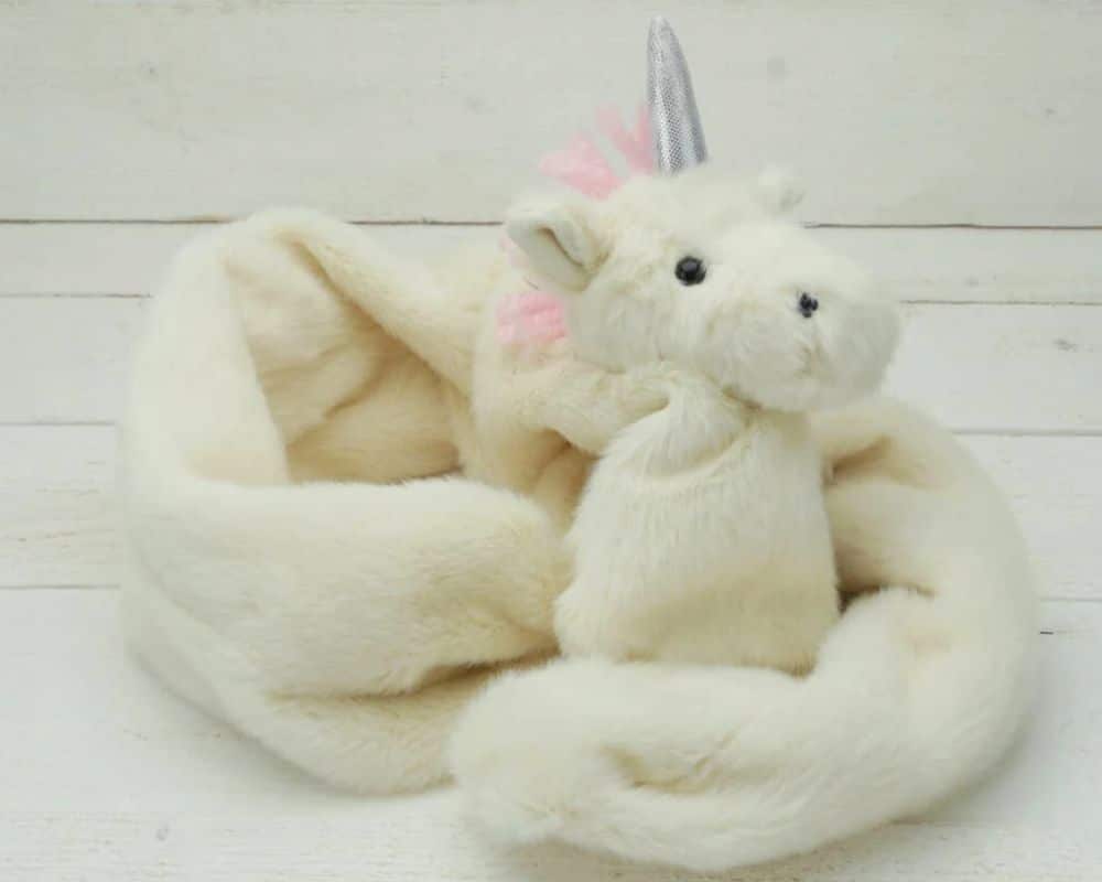 A soft, cream-coloured unicorn scarf made of faux fur, featuring a plush unicorn head with a silver horn, pink mane, and black button eyes, displayed against a white wooden background.