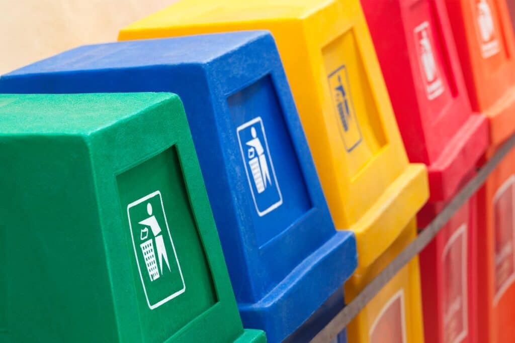 A row of colourful recycling bins—green, blue, yellow, and red—each with a white symbol indicating proper waste disposal, is lined up for tenants to easily sort their recyclables and help keep the community clean.