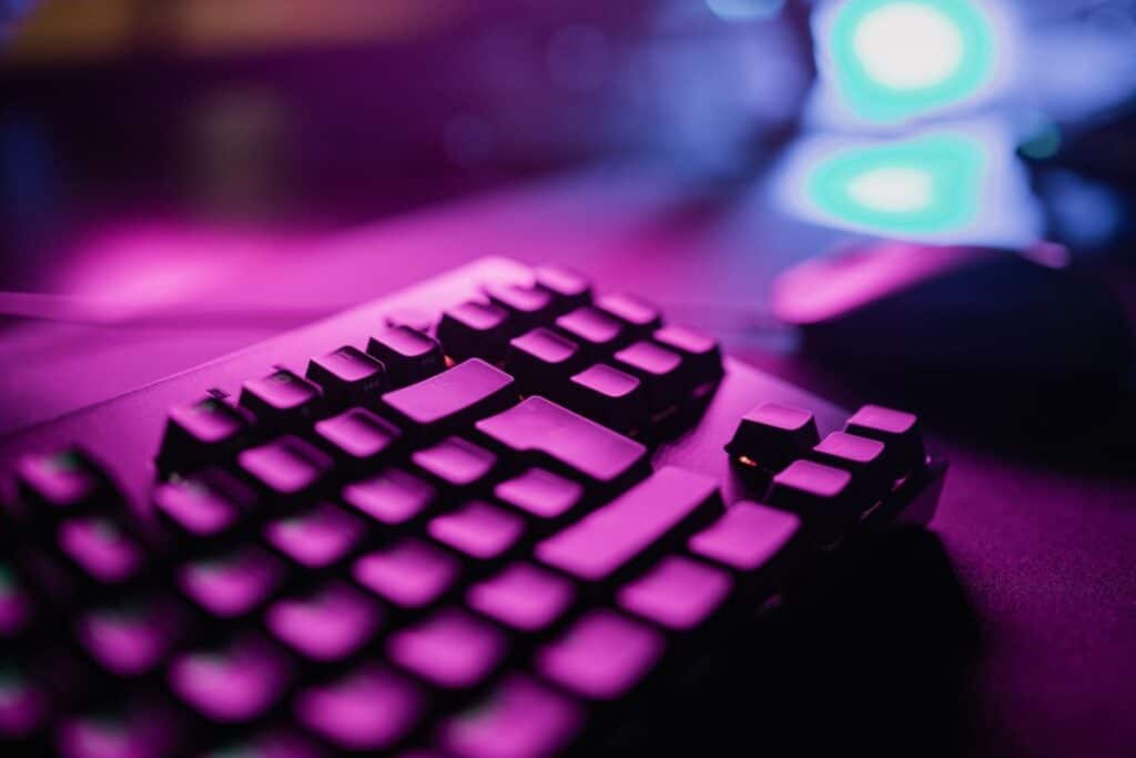 Close-up of a computer keyboard with purple and blue lighting, creating a vibrant and modern gaming atmosphere. A computer mouse is visible in the blurred background.