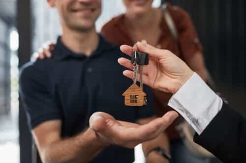 A hand gives house-shaped keys to a smiling couple, suggesting their entry into the housing market as they receive keys to a new home. The couple looks happy and excited.