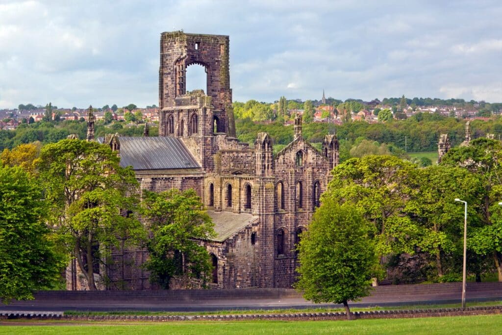 Large, partially ruined stone abbey with a tall, open central tower, surrounded by green trees and a curved road, with new-build homes in the backdrop alongside houses and a cloudy sky.