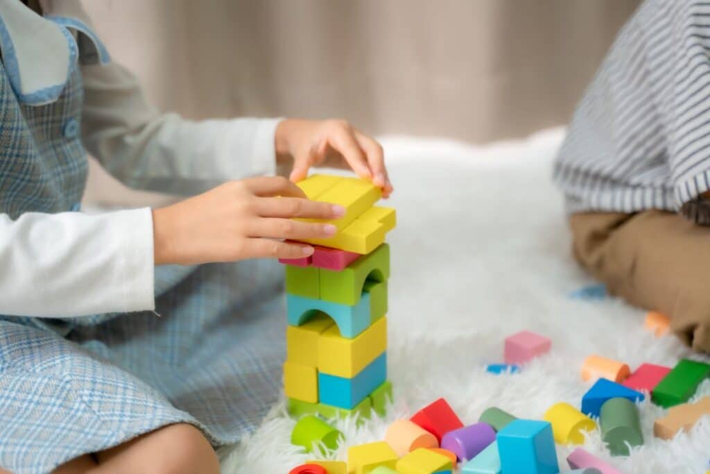 A child builds a colourful tower with wooden blocks on a white, fluffy surface, while another child sits nearby. Various loose toys and blocks are scattered around them.