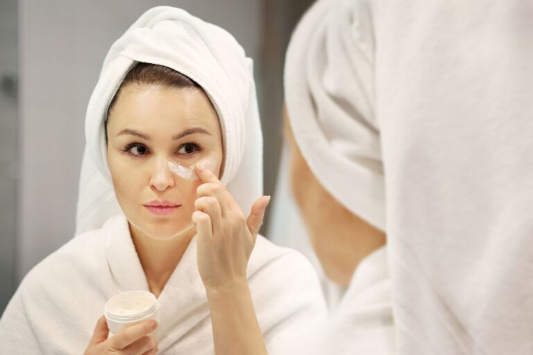 A woman in a white dressing gown and towel wrapped around her head applies anti-ageing cream to her face while looking in a bathroom mirror. She holds a small jar of the cream in her other hand.