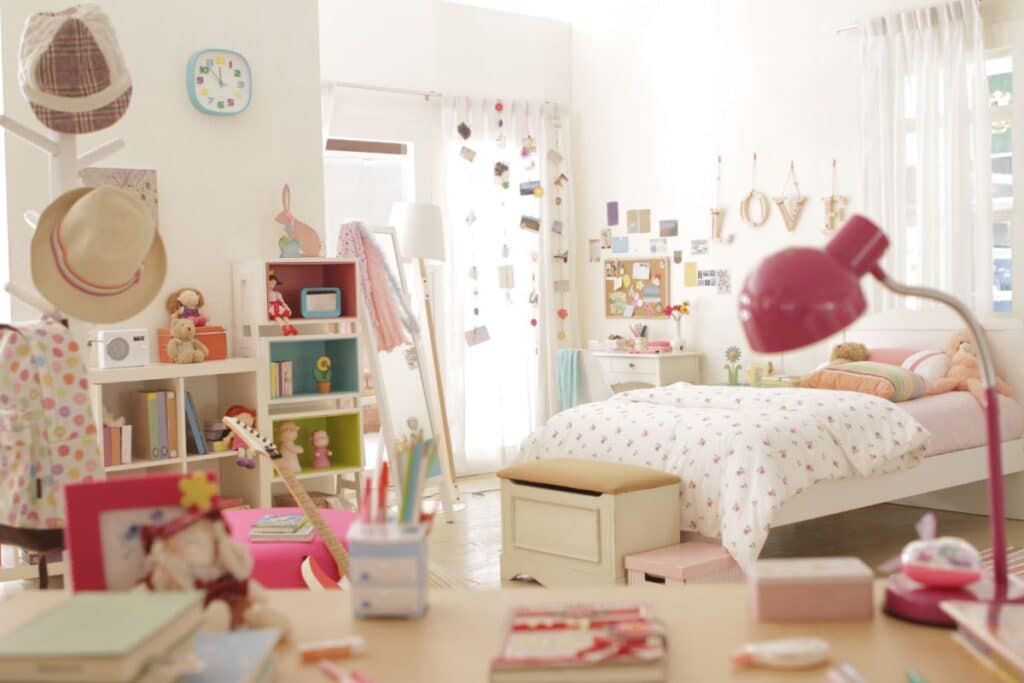 A bright, cosy new bedroom with a neatly made bed, colourful decor, books, and a desk with a pink lamp. Shelves display toys, hats, and photos. Sunlight pours through sheer curtains, creating a cheerful atmosphere.