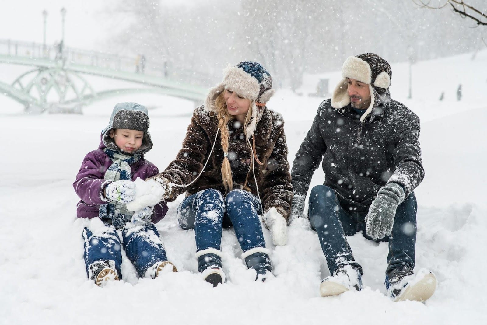 A family of three, dressed in winter clothes and hats, sit together in the snow during a snowfall, enjoying a stress-free moment. A snowy park and a bridge are visible in the background.