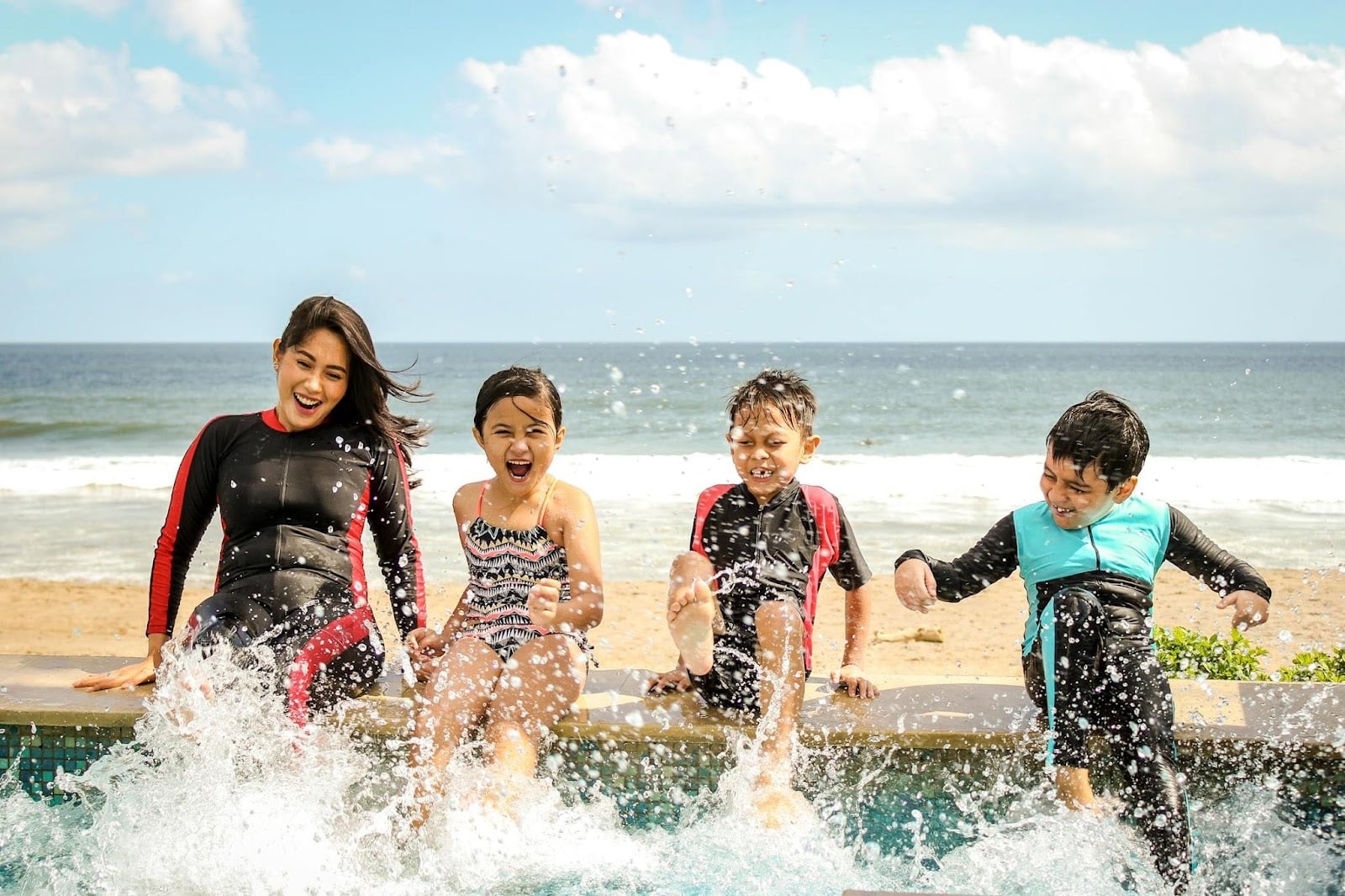 Four children sit on the edge of a pool, laughing and splashing water with their feet under a bright, sunny sky. With the sea and sandy beach behind them, it’s a perfect stress-free moment of carefree fun.