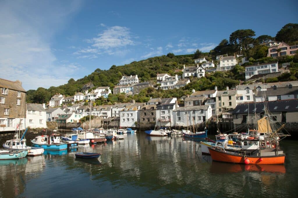 A small, family-friendly harbour with colourful fishing boats moored in calm water, surrounded by quaint white and stone cottages on a hillside covered in green trees under a blue sky.