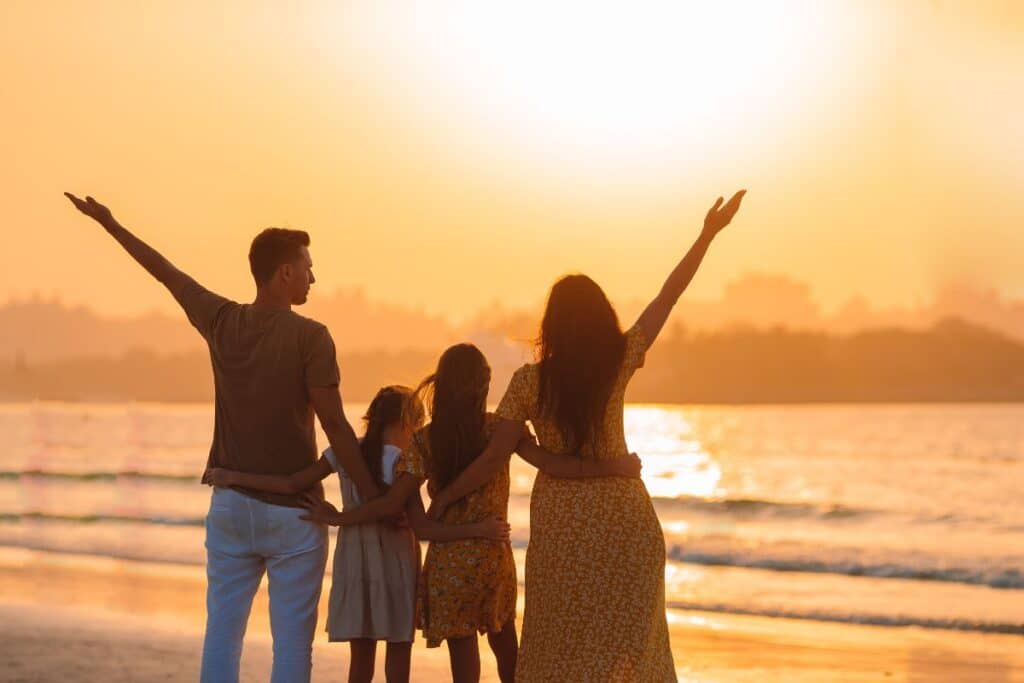 A family-friendly scene unfolds as a family of four stands on the beach at sunset, facing the sea with arms outstretched. The warm sunlight creates a golden glow over the water and silhouettes their figures.