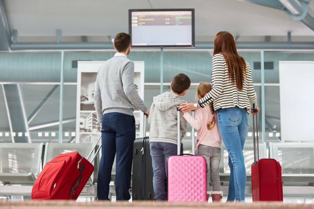 A family of four with suitcases stands in an airport, looking up at a digital departures board. The parents, son, and daughter are seen from behind, waiting in a modern terminal, their posture showing a hint of travel stress.