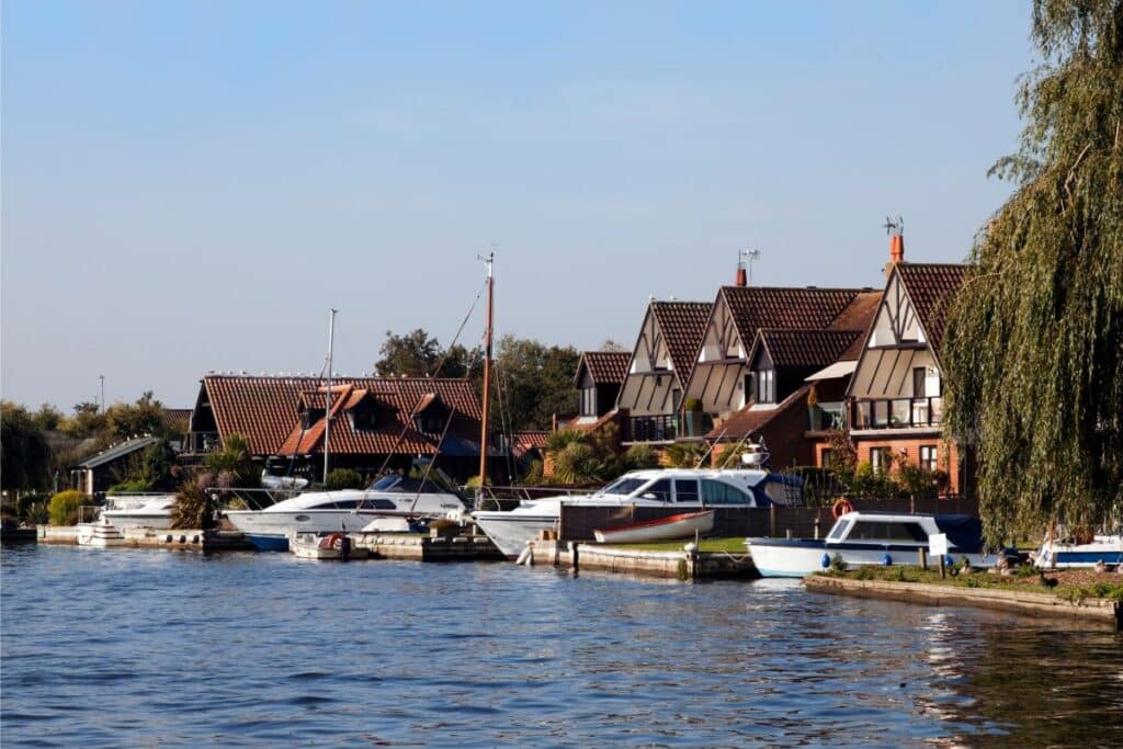 Waterside houses with sloping roofs and timber frames line the Norfolk riverbank, with several boats moored along the edge. A tree overhangs the water on the right, and the sky is clear and blue.