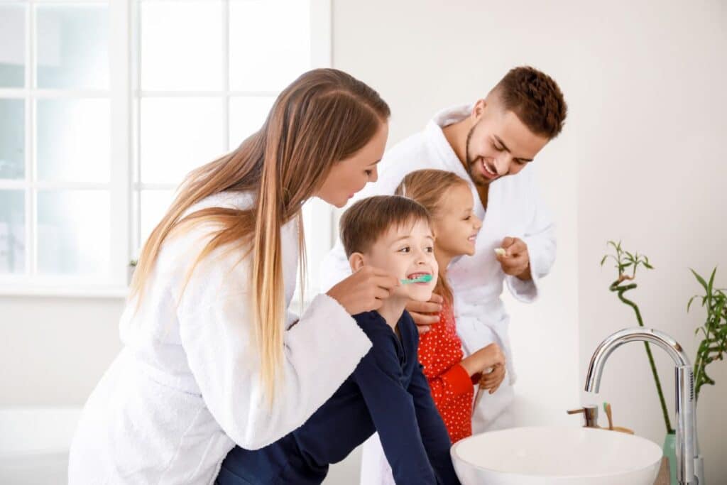 A family of four in dressing gowns and pyjamas practises oral hygiene together, smiling as they brush their teeth around a bright bathroom basin.