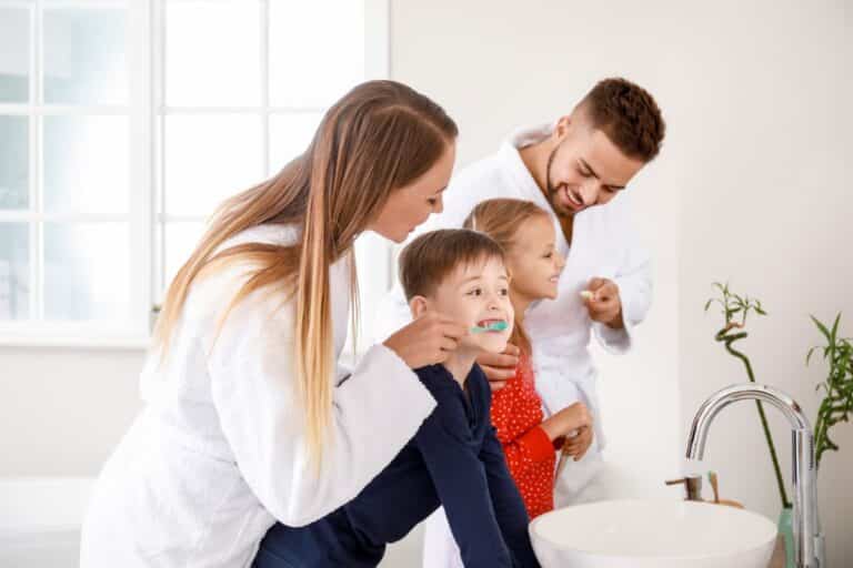 A family of four in dressing gowns and pyjamas practises oral hygiene together, smiling as they brush their teeth around a bright bathroom basin.